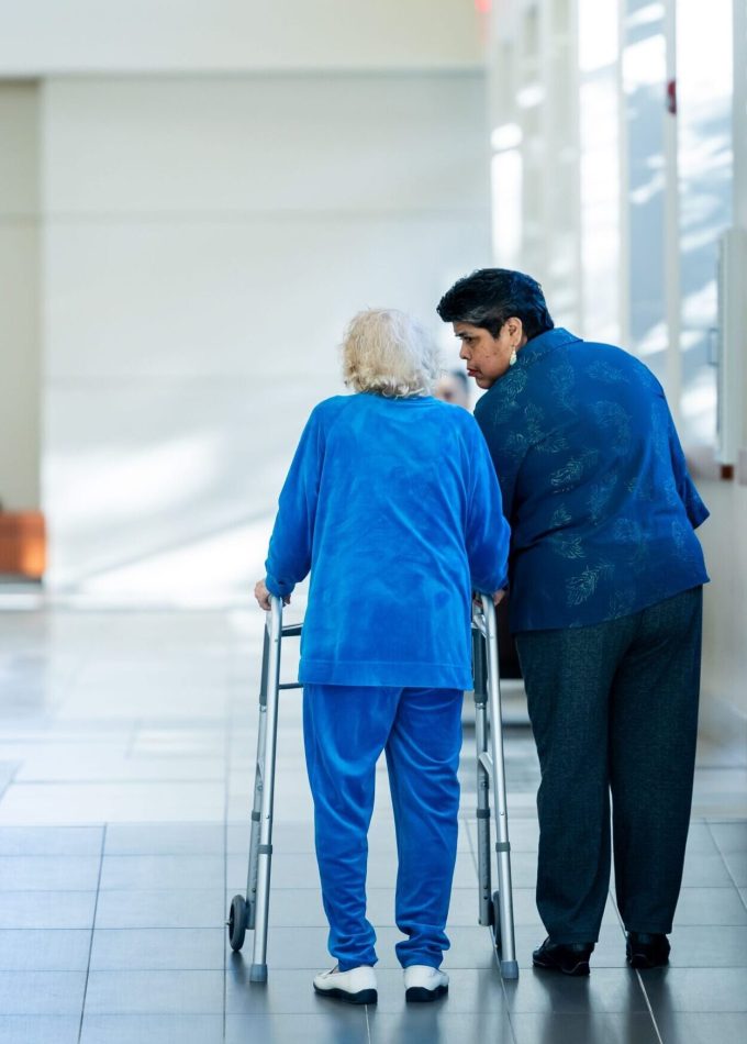 A nurse assists an elderly woman with a walker in a bright hospital corridor.